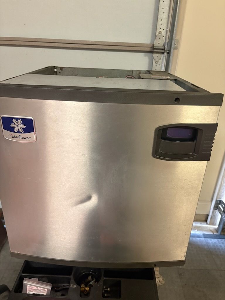 Stainless steel commercial ice maker with black dispenser panel in a utility room.