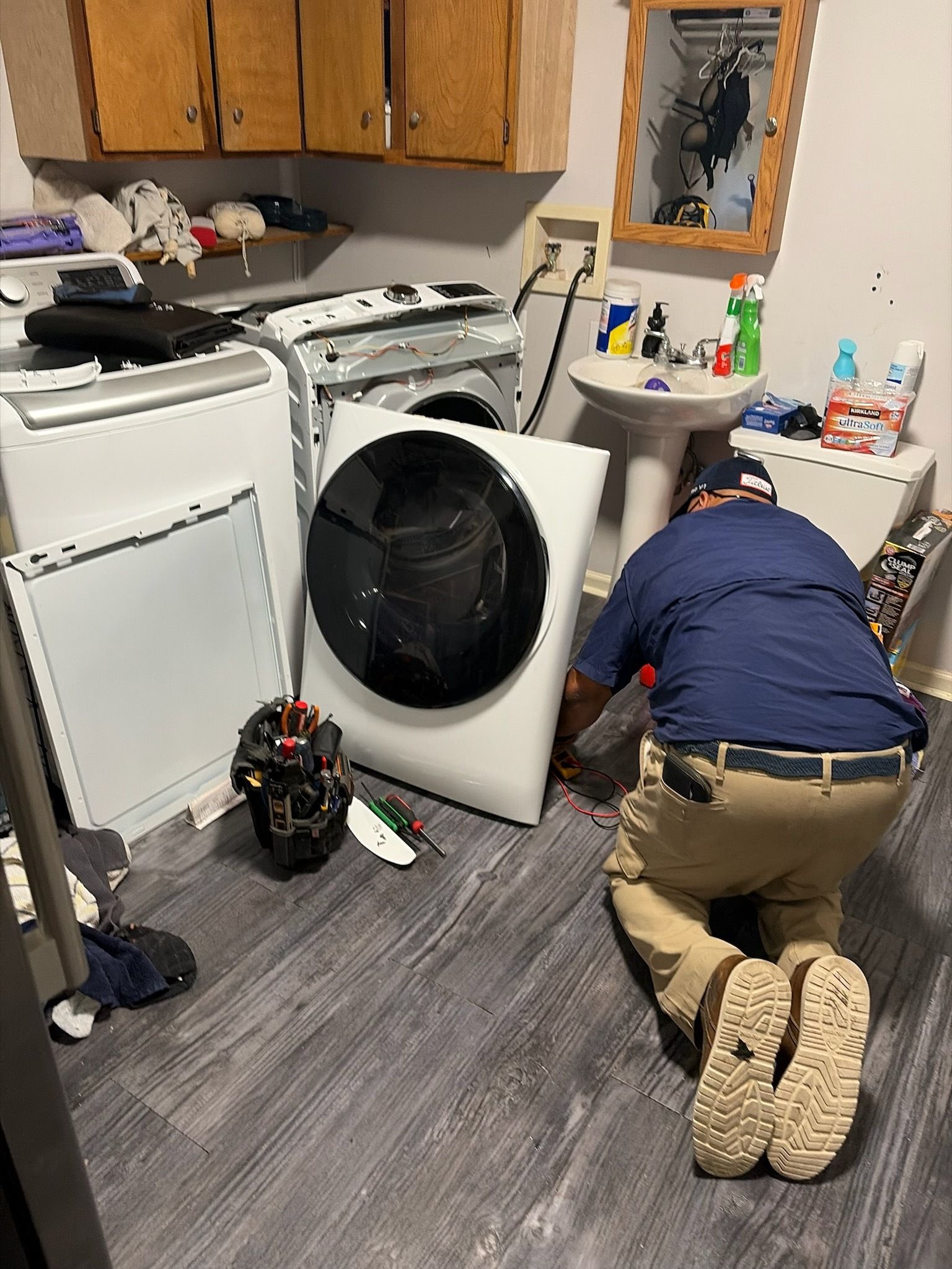 Person kneeling beside a front-loading washer in a cluttered laundry room, facing a cabinet.