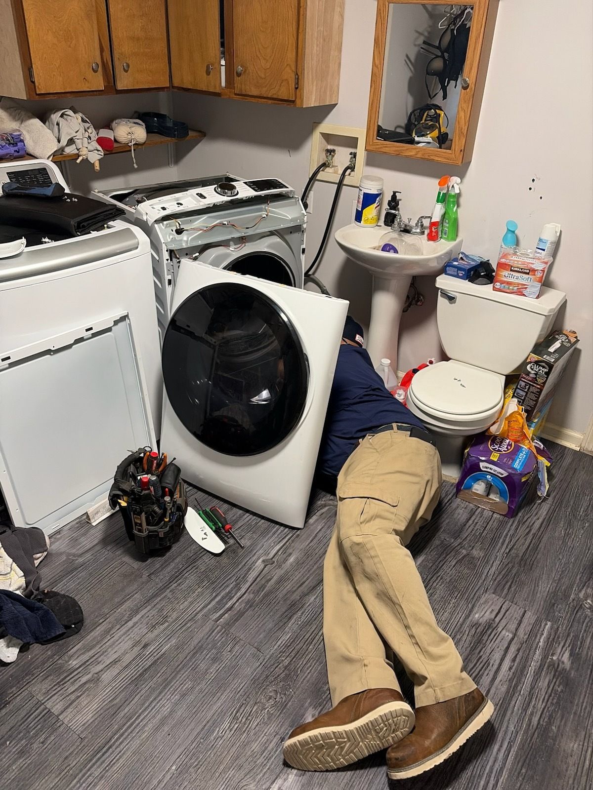 Messy laundry room with washer, dryer, sink, scattered items, and a person lying on the floor