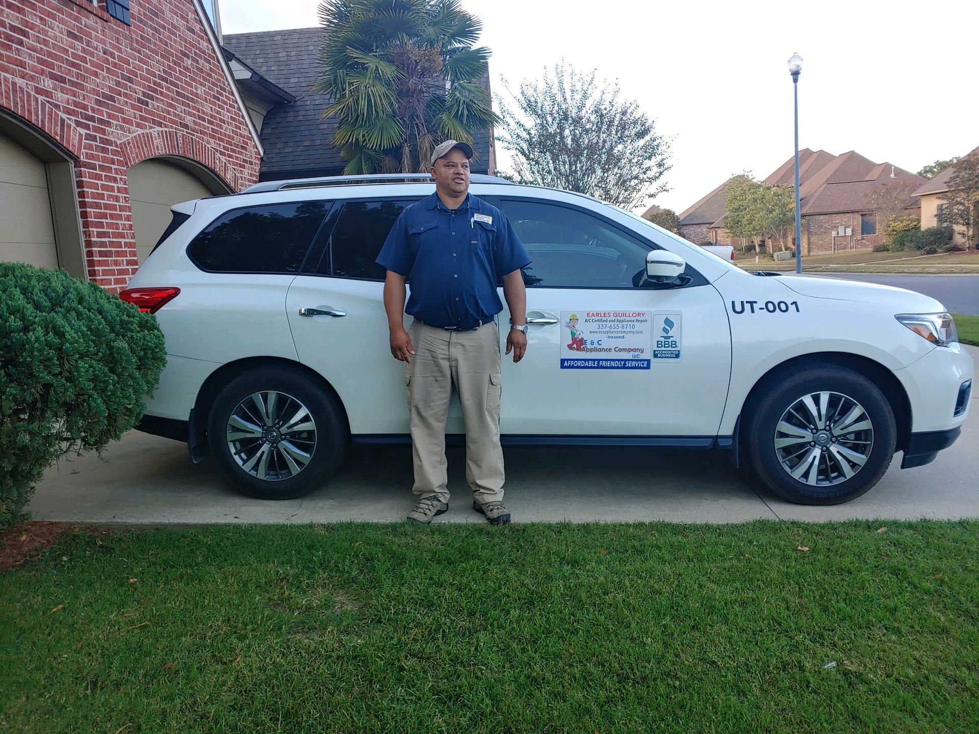 Man standing beside a white SUV in a suburban driveway with a lawn and brick houses.