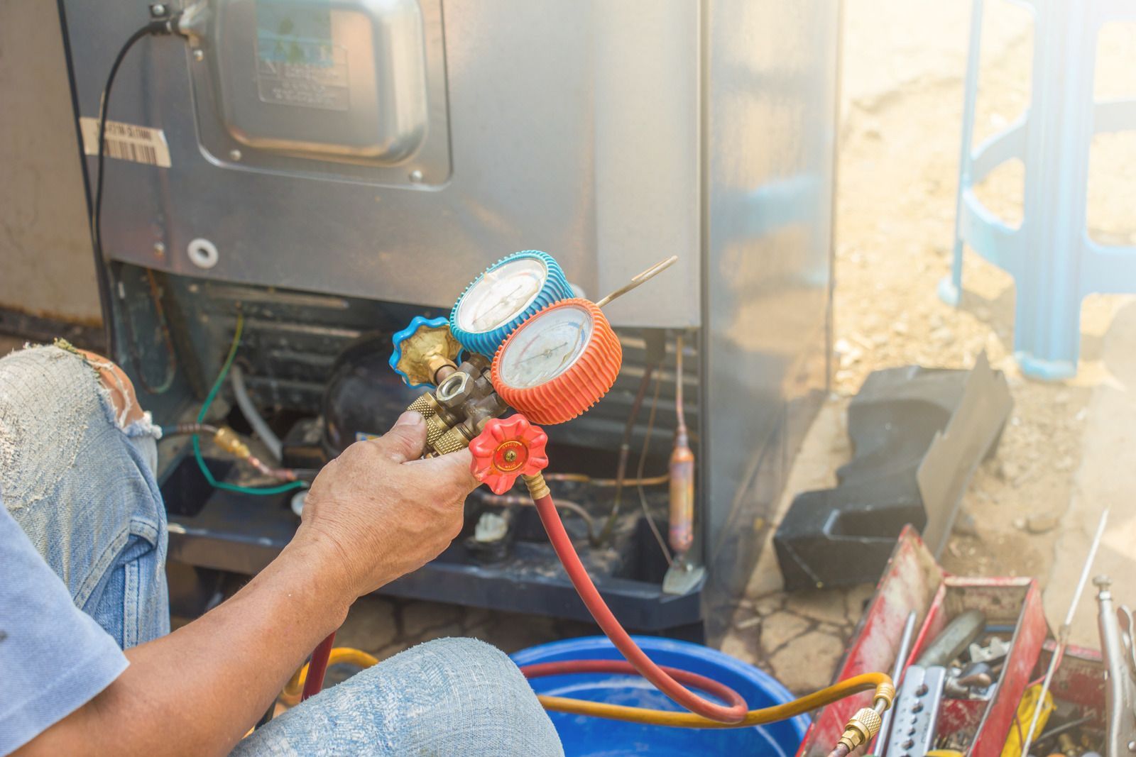 Technician holding HVAC manifold gauges beside an outdoor unit, with blue and red hoses attached
