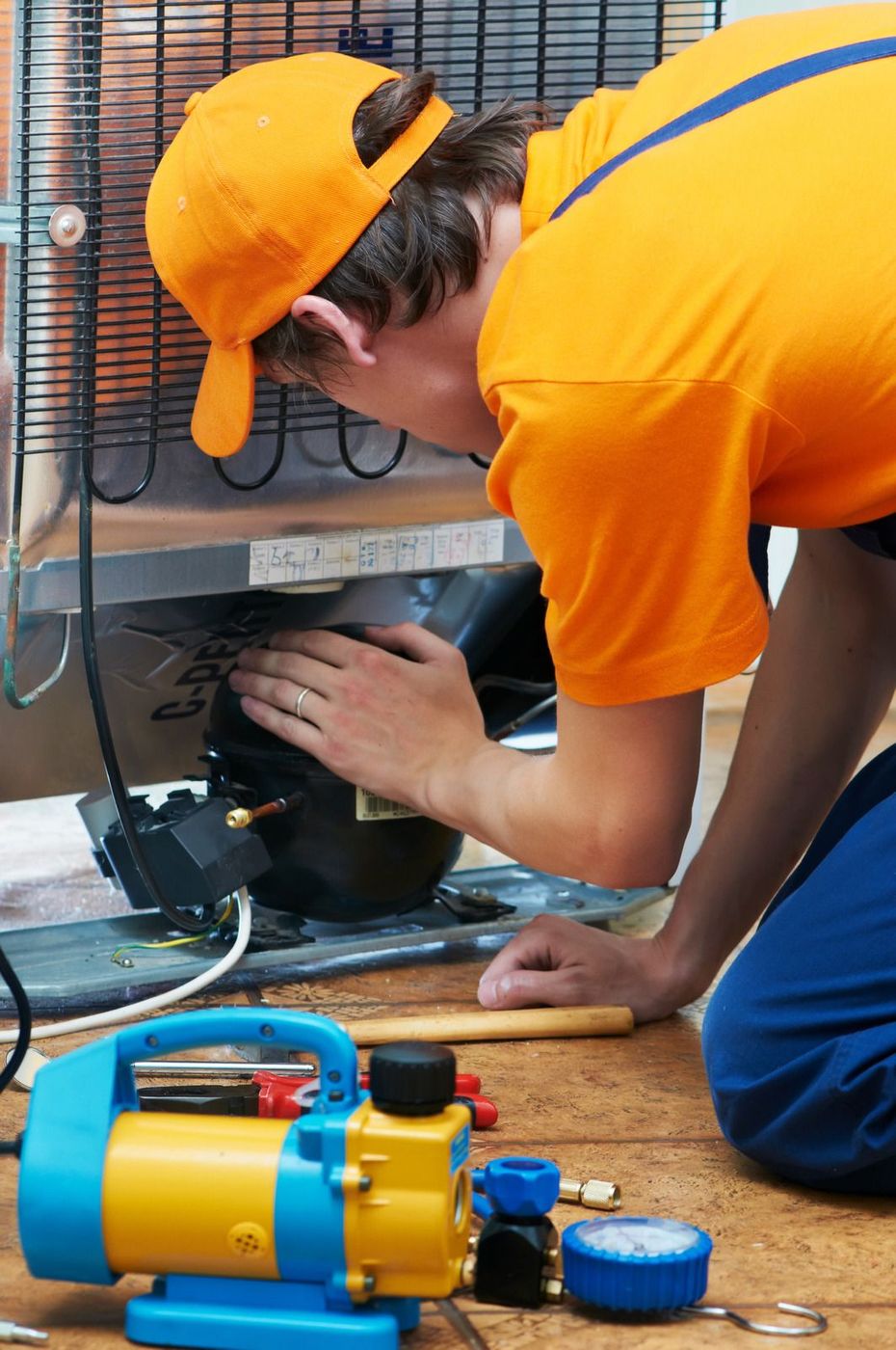 Technician in orange uniform adjusting equipment beneath a workbench beside a yellow air compressor.