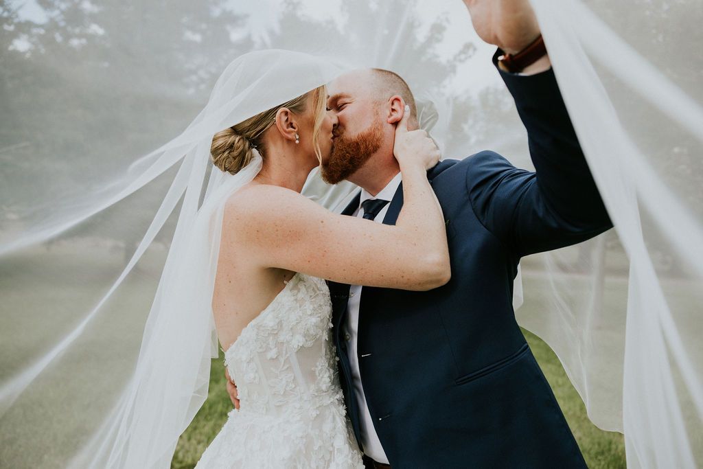 A bride and groom in a dark suit kiss under a sheer wedding veil held up against a blurred green background.