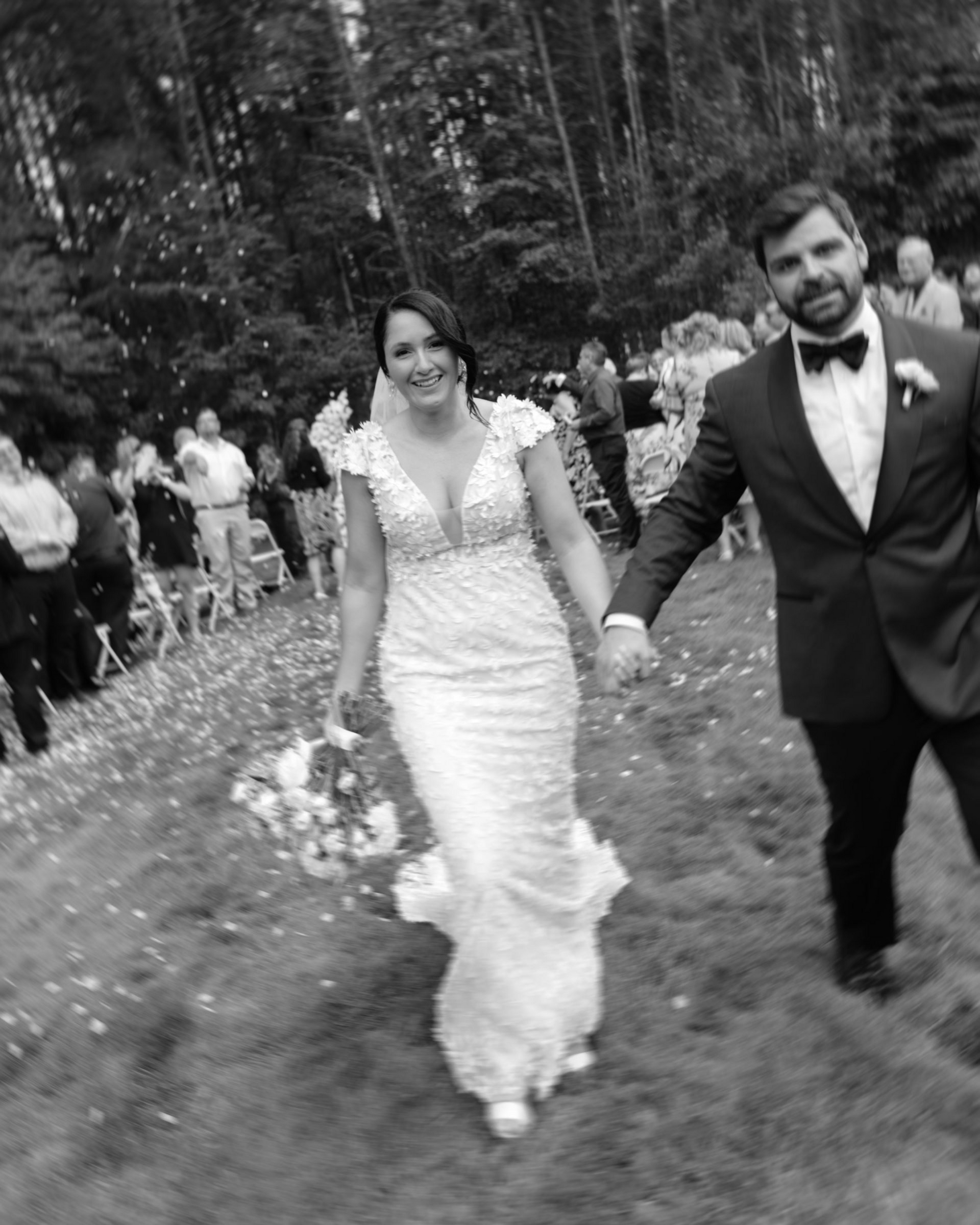 A bride and groom walk hand-in-hand through a grassy outdoor area, smiling as guests cheer in the background.