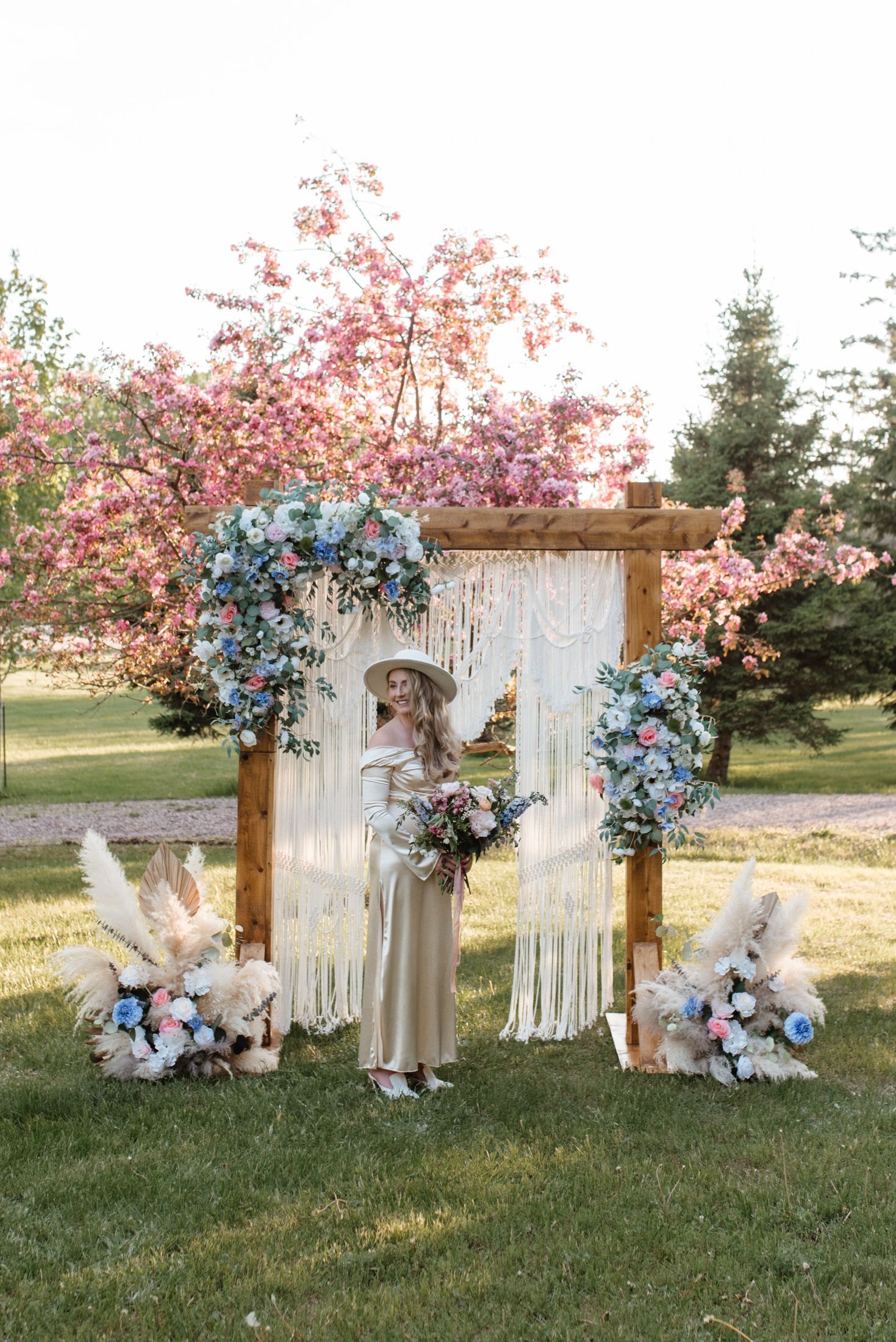 A person in a bohemian dress and hat stands under a floral-adorned wooden arch with macramé accents in a spring garden.