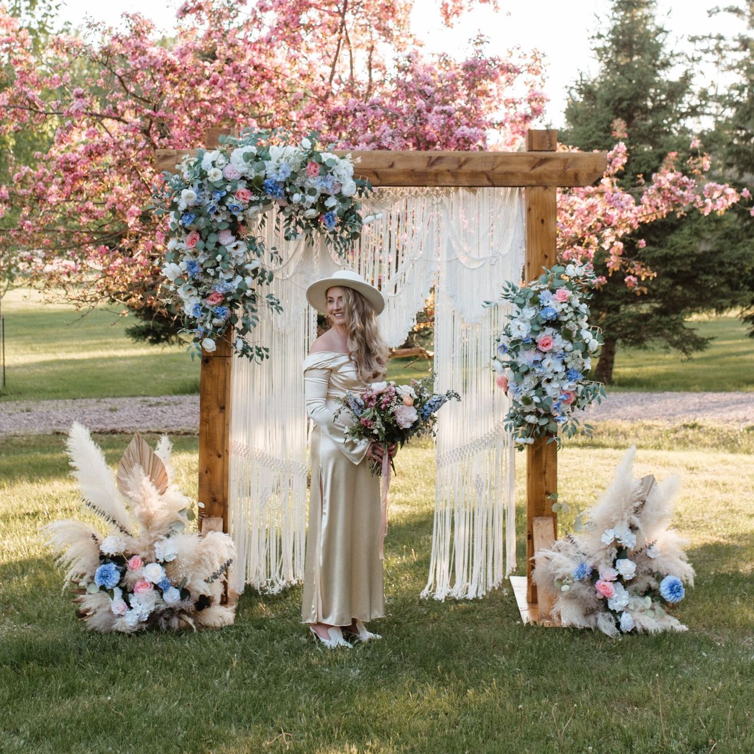 A person in a cream-colored dress and hat stands before a macramé wedding arch decorated with pink and blue floral sprays.