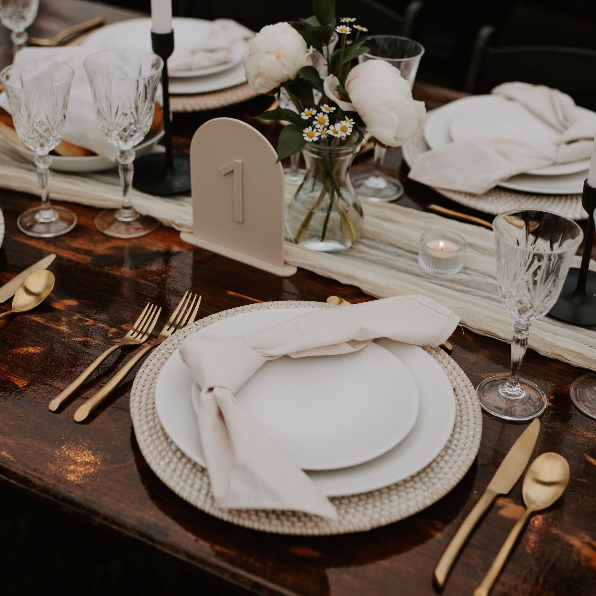 A rustic wedding table setting with white plates, napkins, gold cutlery, crystal glasses, and a numbered table card.