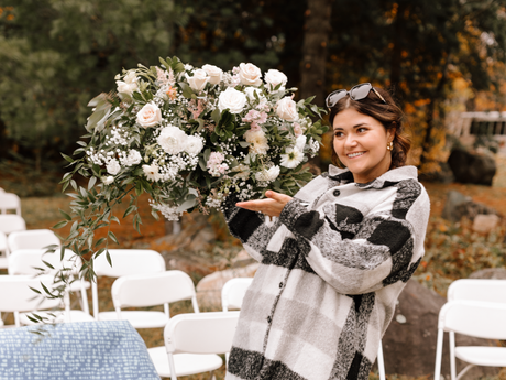 Sarah from Tickled Pink Brides smiling person in a plaid shirt gestures toward a large, white floral arrangement
