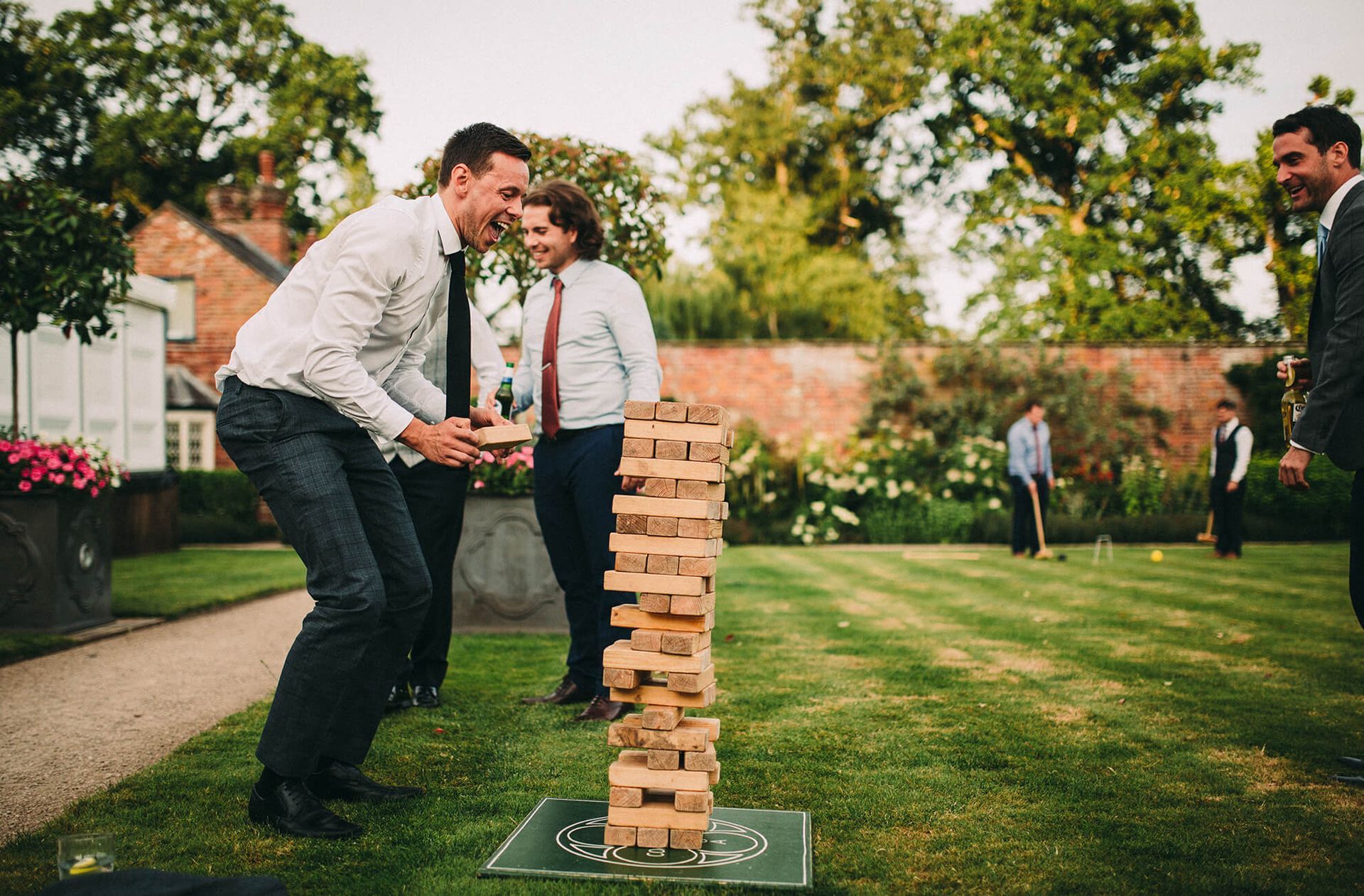 Men in dress clothes play a giant wooden block tower game on a sunny lawn at an outdoor event.