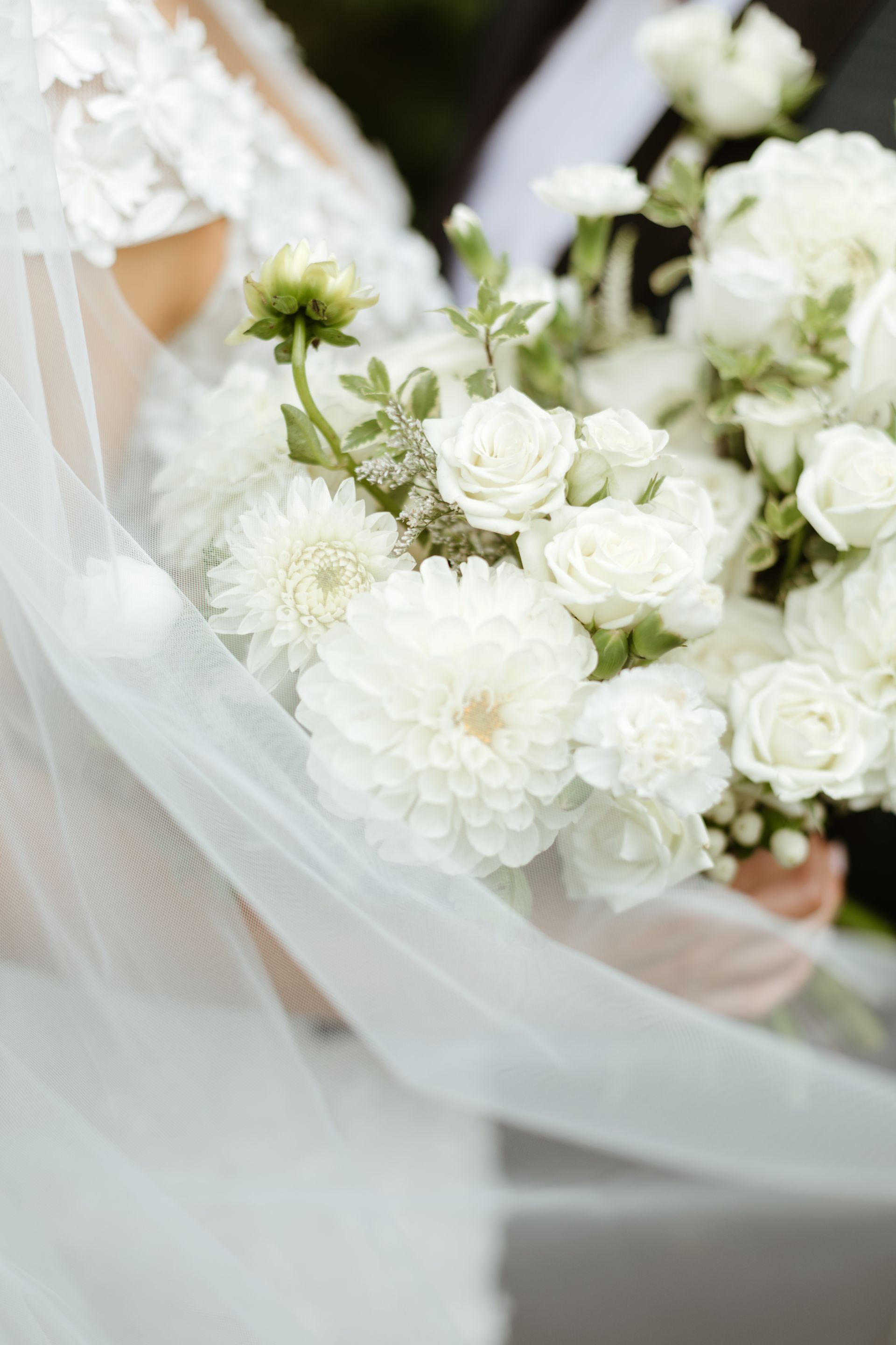 A close-up of a bride holding a white bouquet of dahlias, roses, and greenery, partially covered by a flowing white veil.