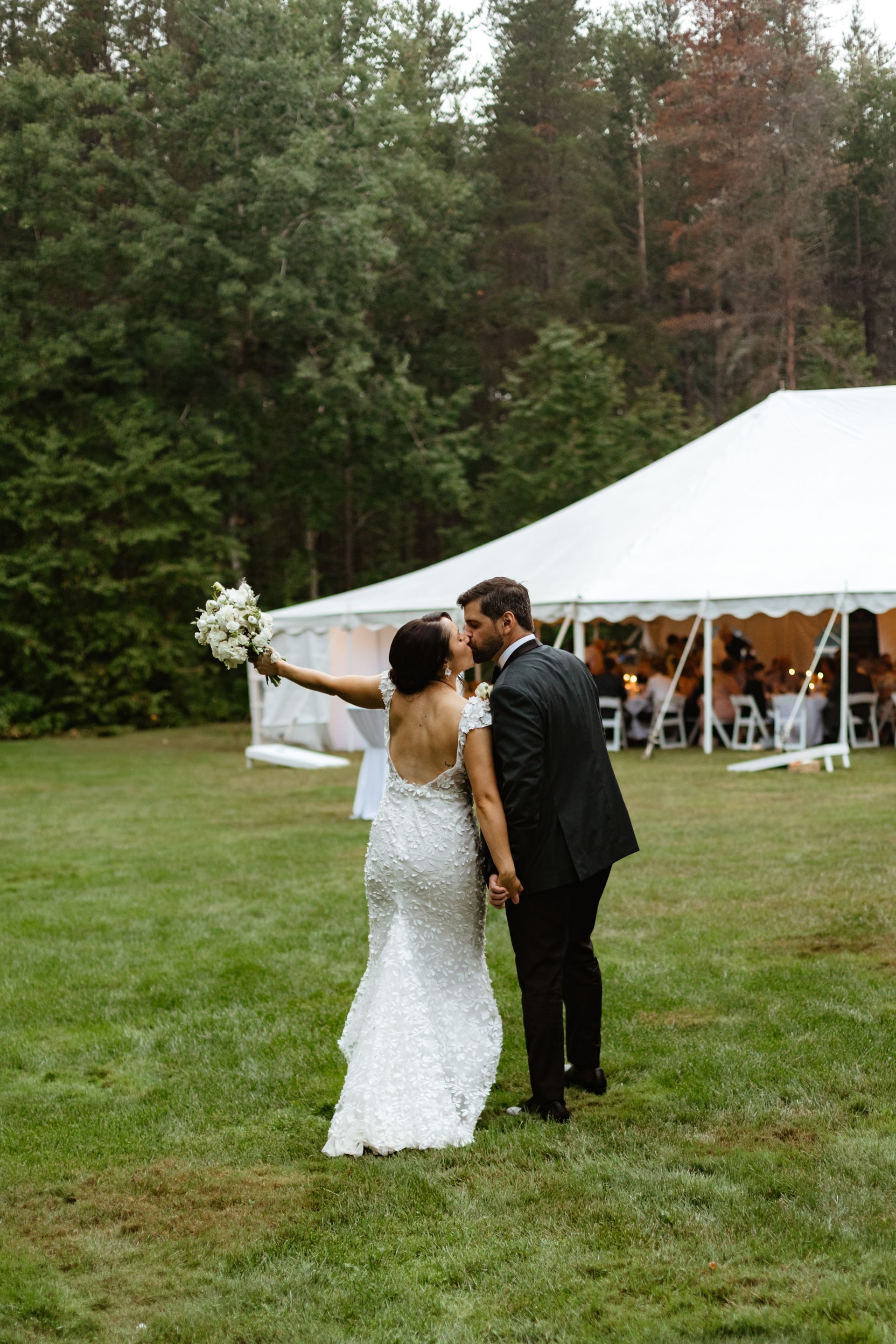 A bride and groom in wedding attire kiss while walking on a grassy lawn toward a white event tent in a wooded area.