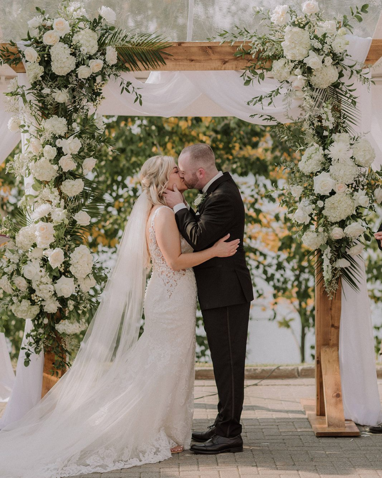 A newlywed couple kisses under a wooden wedding arch decorated with white flowers and greenery on a paved patio.