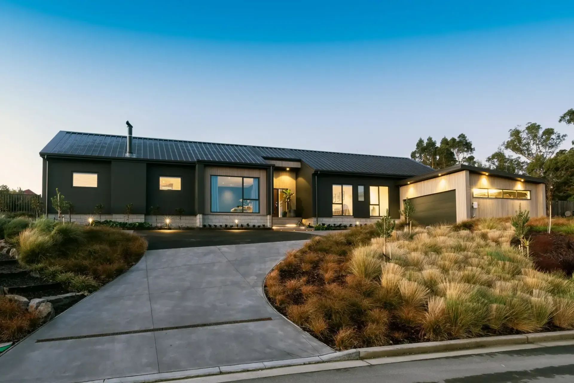 Modern dark-colored house with concrete driveway, landscaping, and garage under a blue sky.