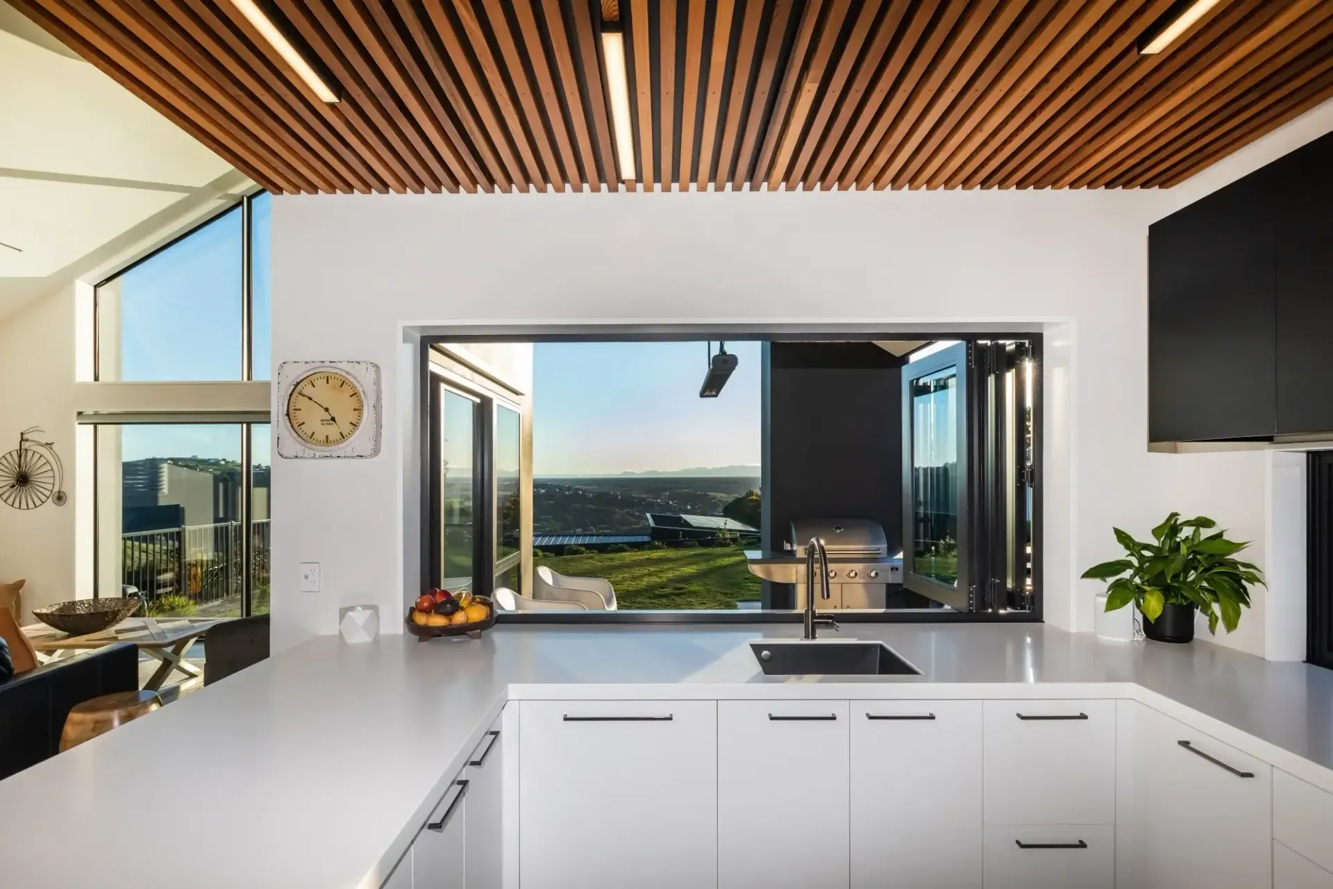 Modern kitchen with folding windows, white cabinets, and wood ceiling, overlooking a scenic landscape.