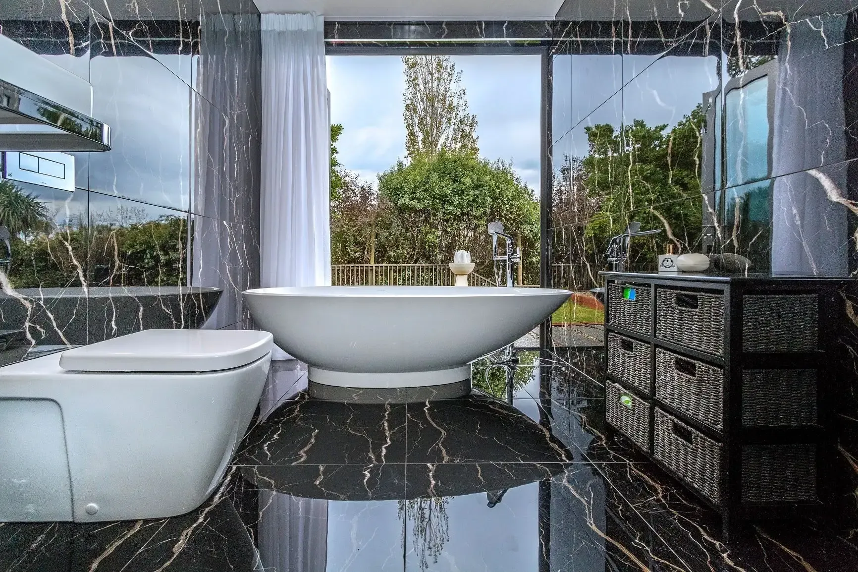 Modern bathroom with black marble, white bathtub, and large window overlooking a garden.