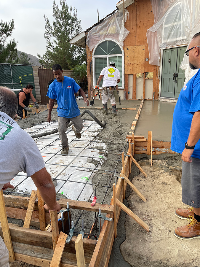 Construction workers pouring concrete pathway at a house. Workers in blue shirts, wooden forms, cement truck.