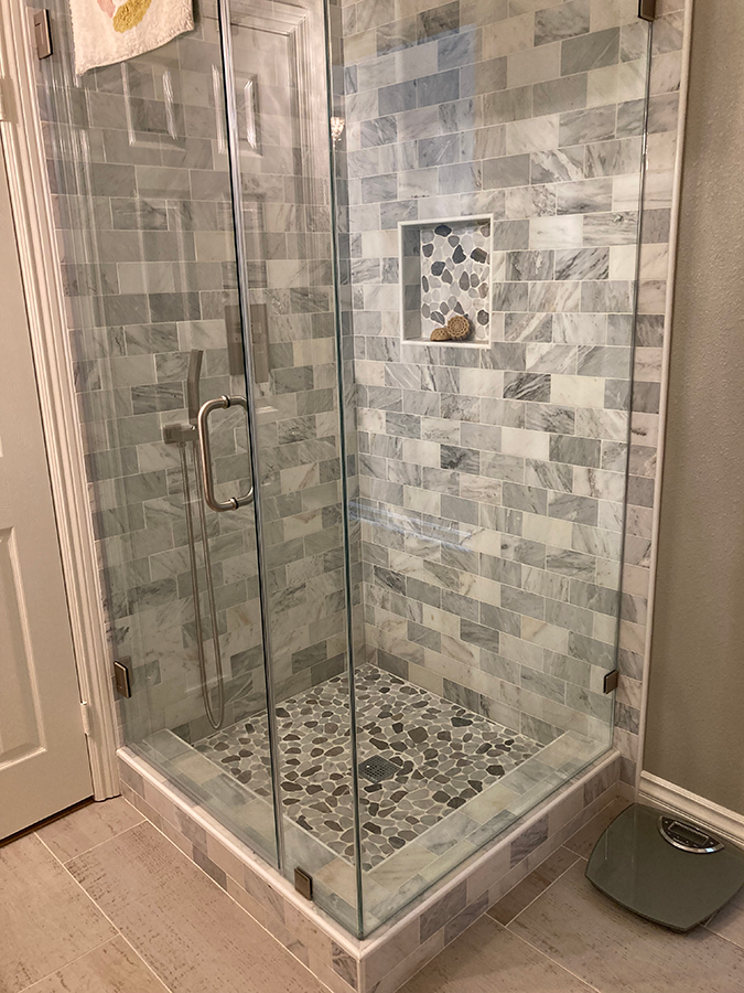 Glass-enclosed shower with gray marble tile, pebble floor, and built-in shelf.