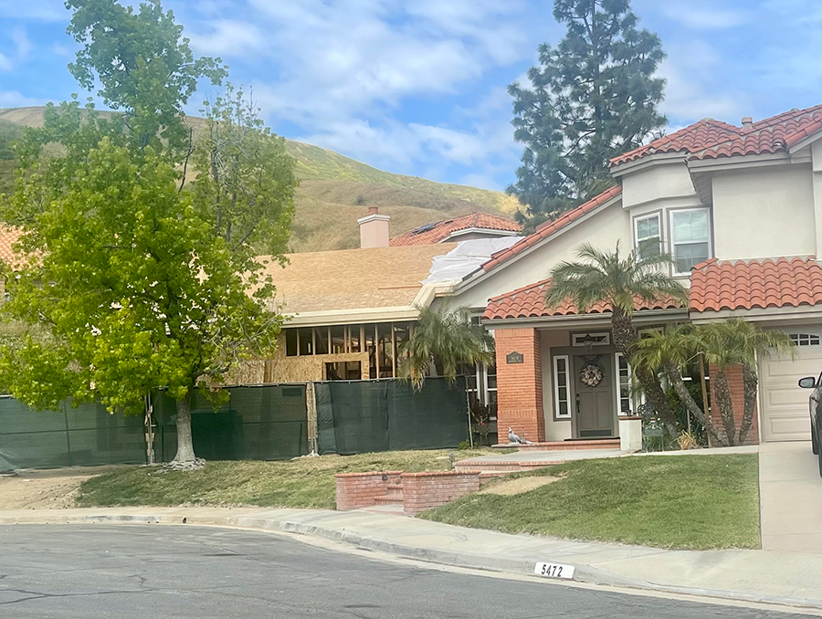 Houses in a suburban neighborhood; one under construction behind a green fence, the other with red tile roof and palm trees.