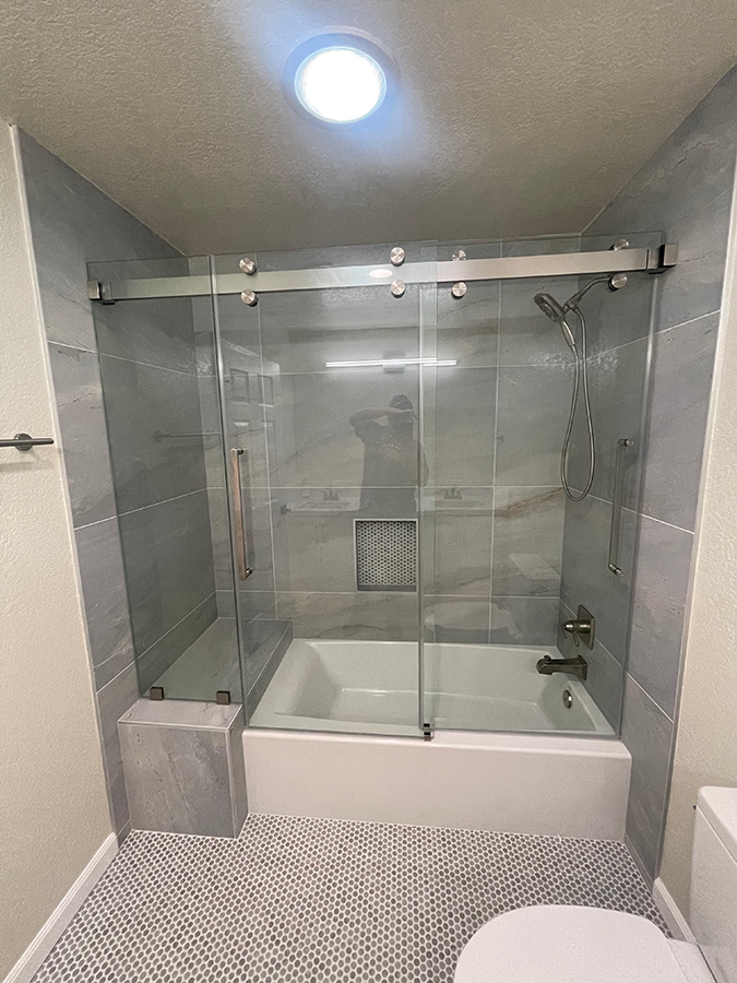 Bathroom with glass shower doors and white tub; gray tile walls and floor; toilet on right.