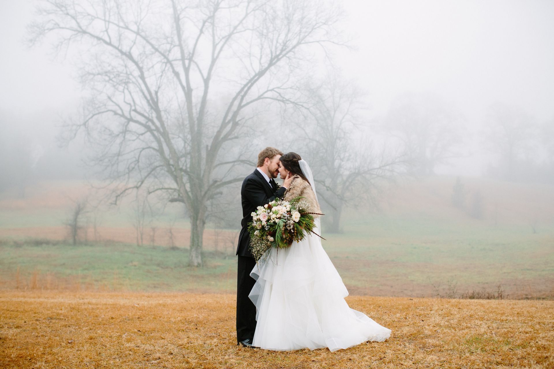 Bride and groom embrace in a foggy field with autumn leaves and bare trees