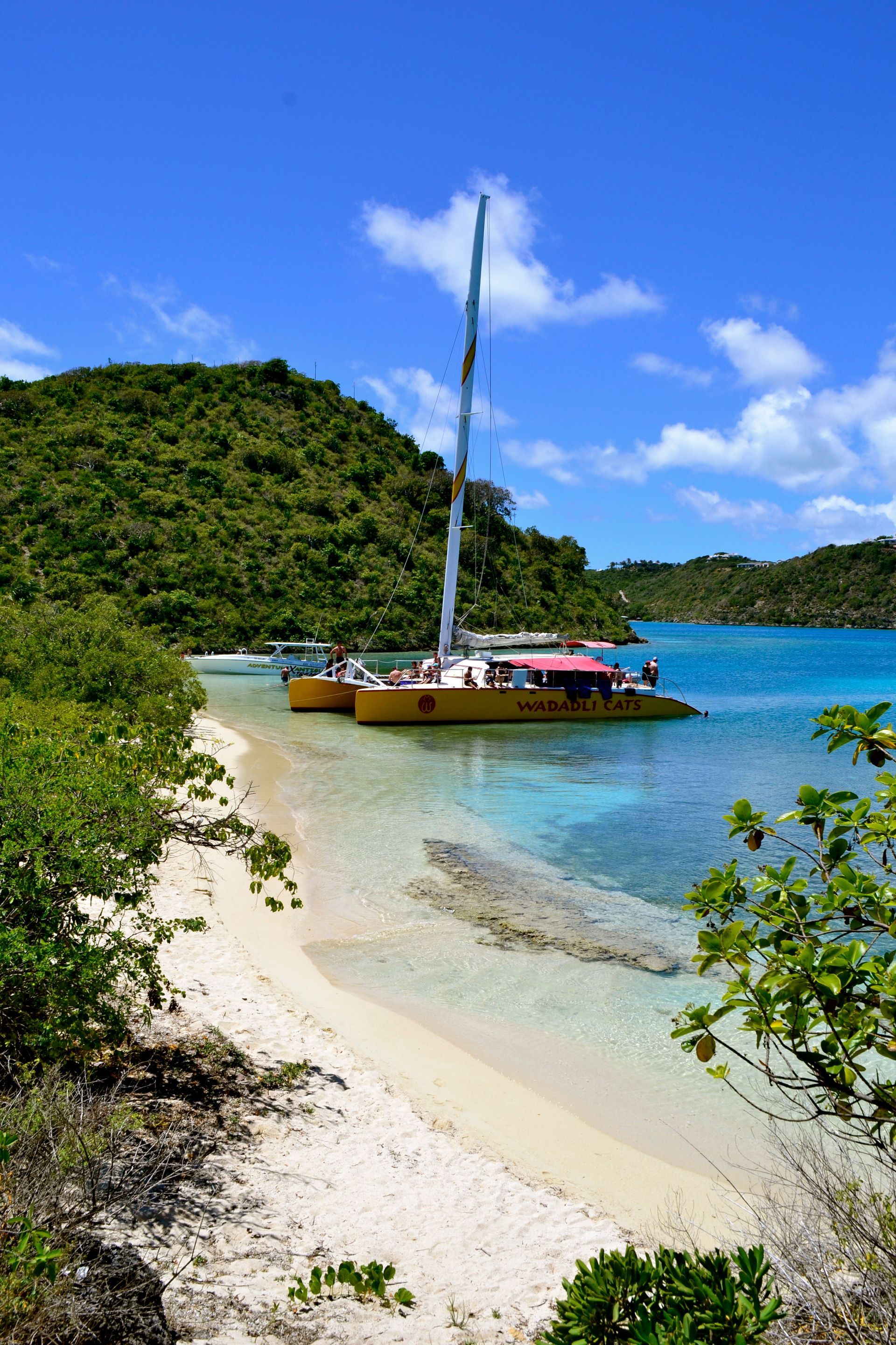 Sailboat anchored in a turquoise tropical cove beside a sandy beach and green hills under a blue sky