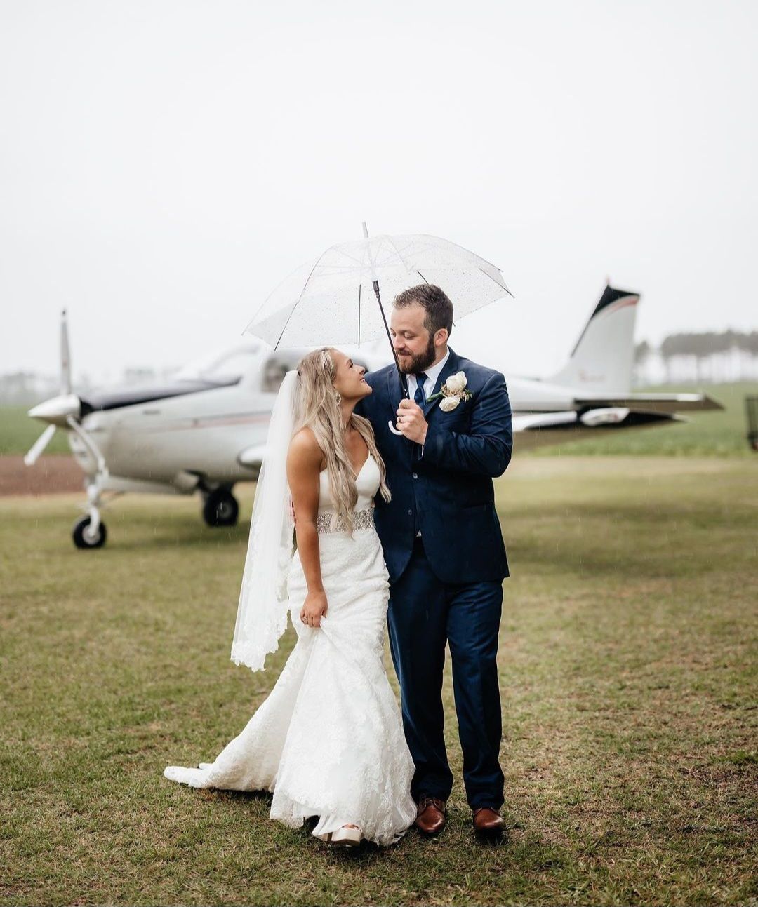 bride and groom smiling under clear umbrella in front of plane