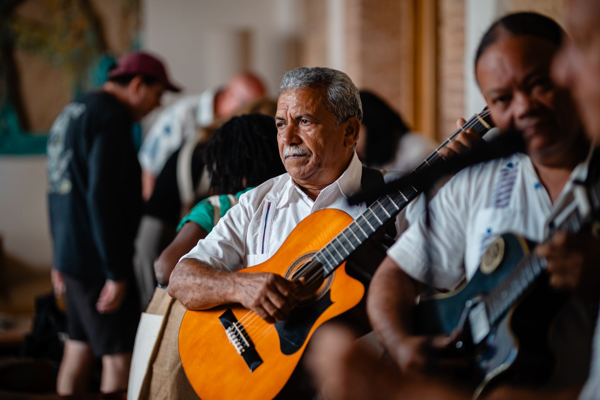 Musicians playing guitars indoors, with a man in a white shirt centered in the foreground