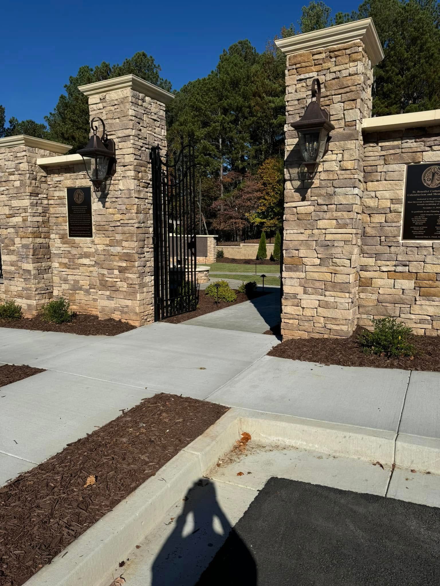 Stone gate entrance with open iron gate, brick pillars, and trees beyond a sidewalk.