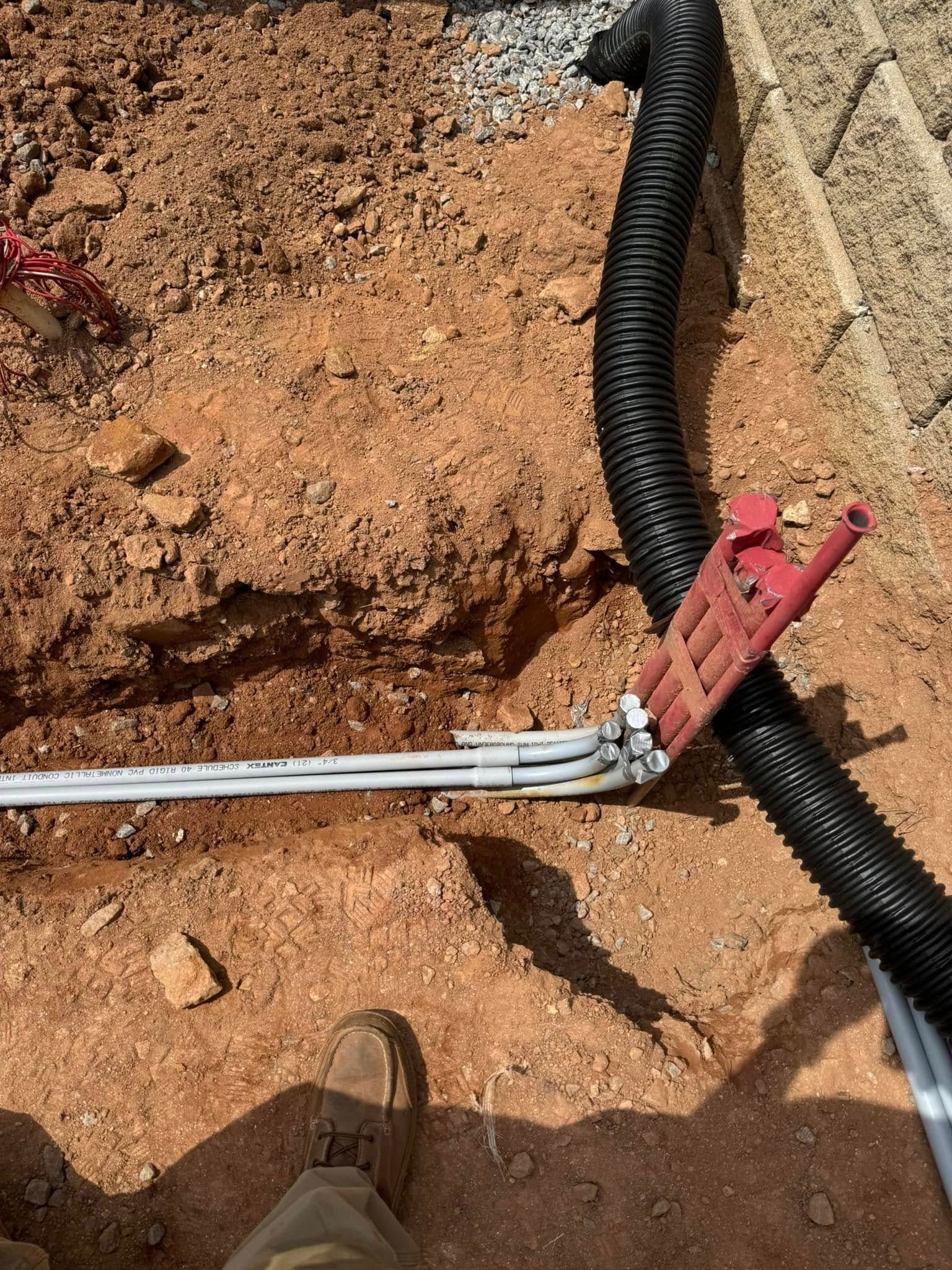 Trench with exposed utility conduit and black corrugated pipe beside a concrete curb and red clamp
