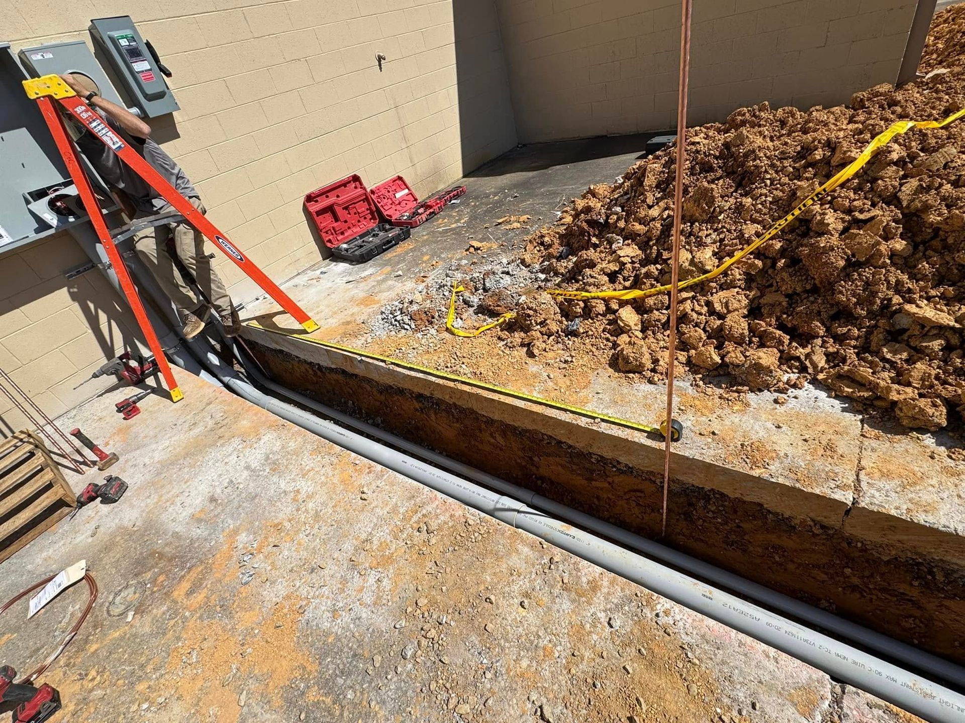 Trench along a concrete wall with black pipe, dirt pile, and tools at a construction site.