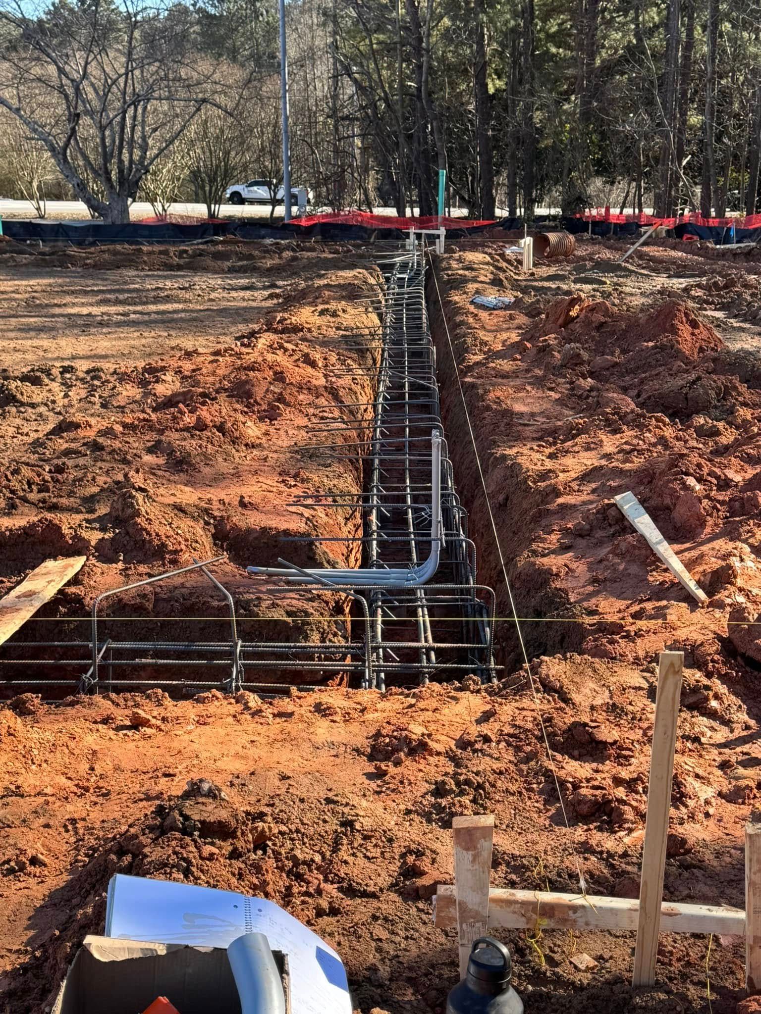 Construction site with trench and exposed rebar foundation, bordered by orange dirt and trees in the background