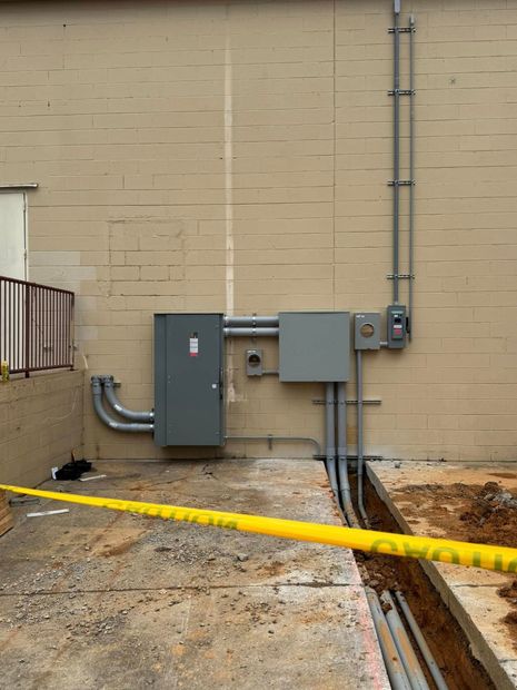 Side of a building with electrical boxes, conduit, and an open trench marked by yellow caution tape.
