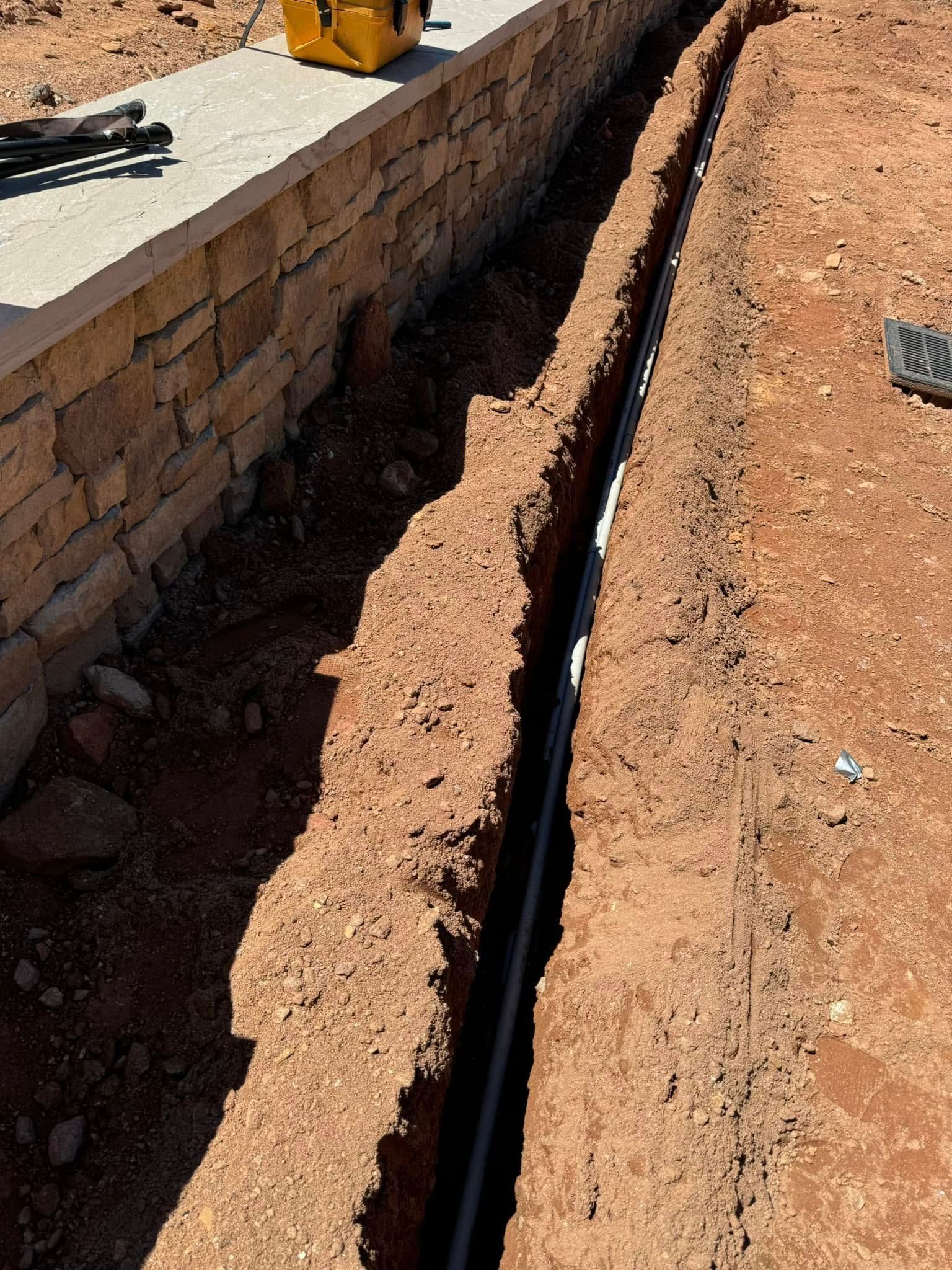 Trench with black pipe running beside a brick retaining wall on a dirt construction site