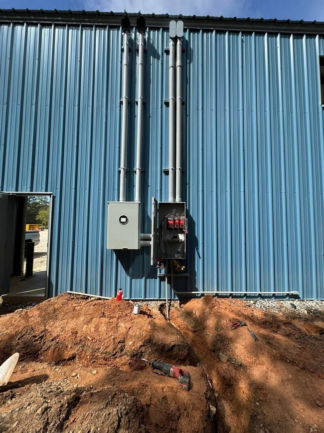 Blue corrugated metal building exterior with electrical panels and conduit above a dirt trench