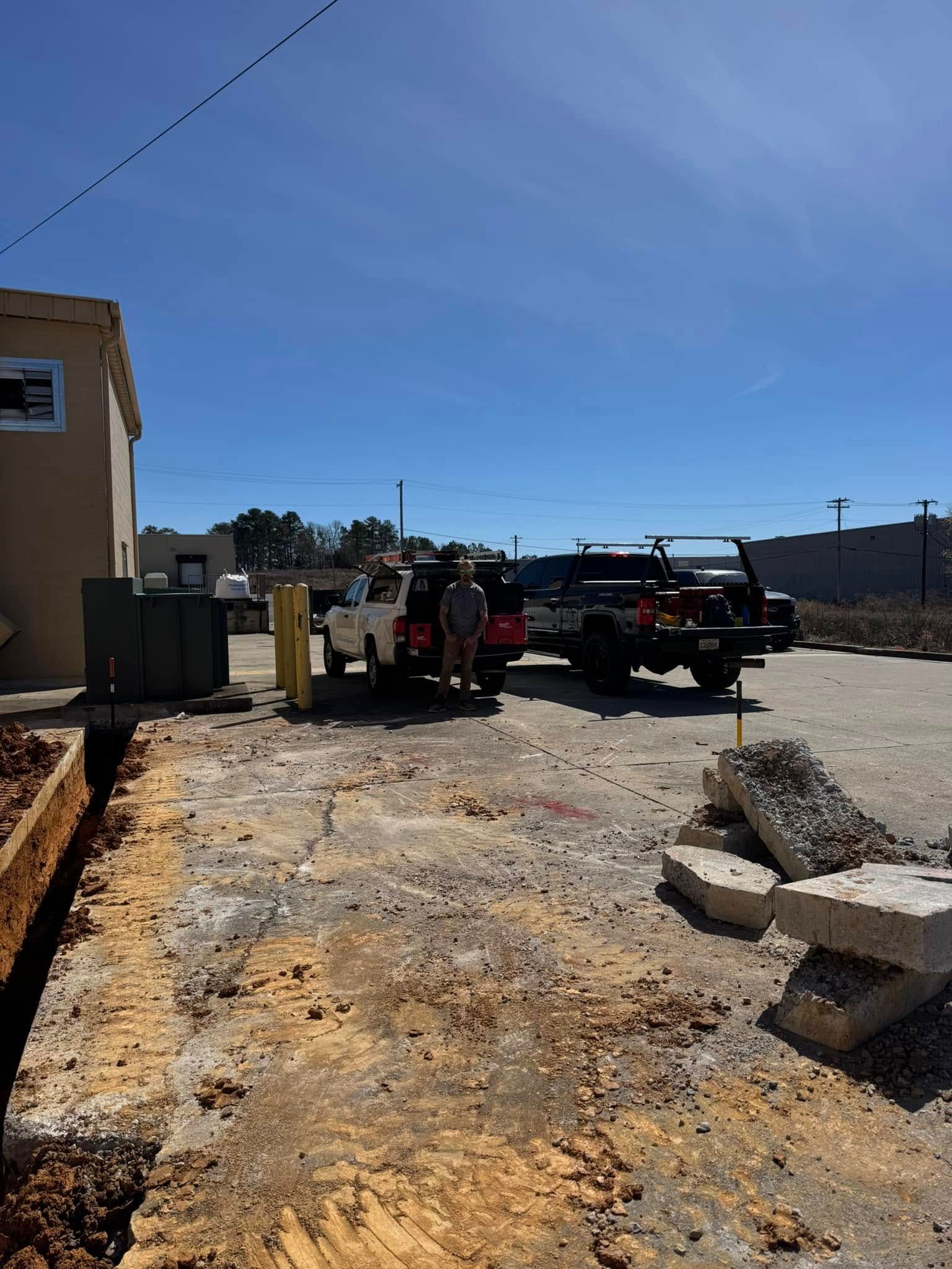Sandy construction lot beside a beige building, with a work truck and equipment under a clear blue sky.