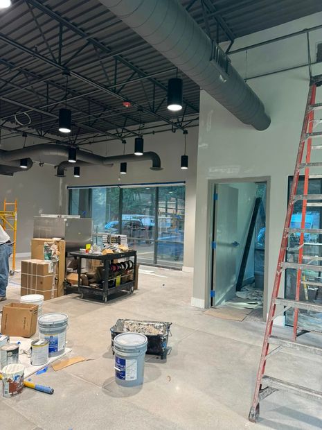 Construction site interior with ladders, boxes, paint buckets, and large windows under exposed ceiling lights