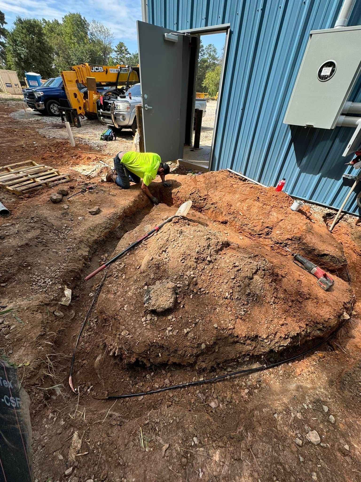 Worker in neon vest digging beside a blue metal building at a construction site.