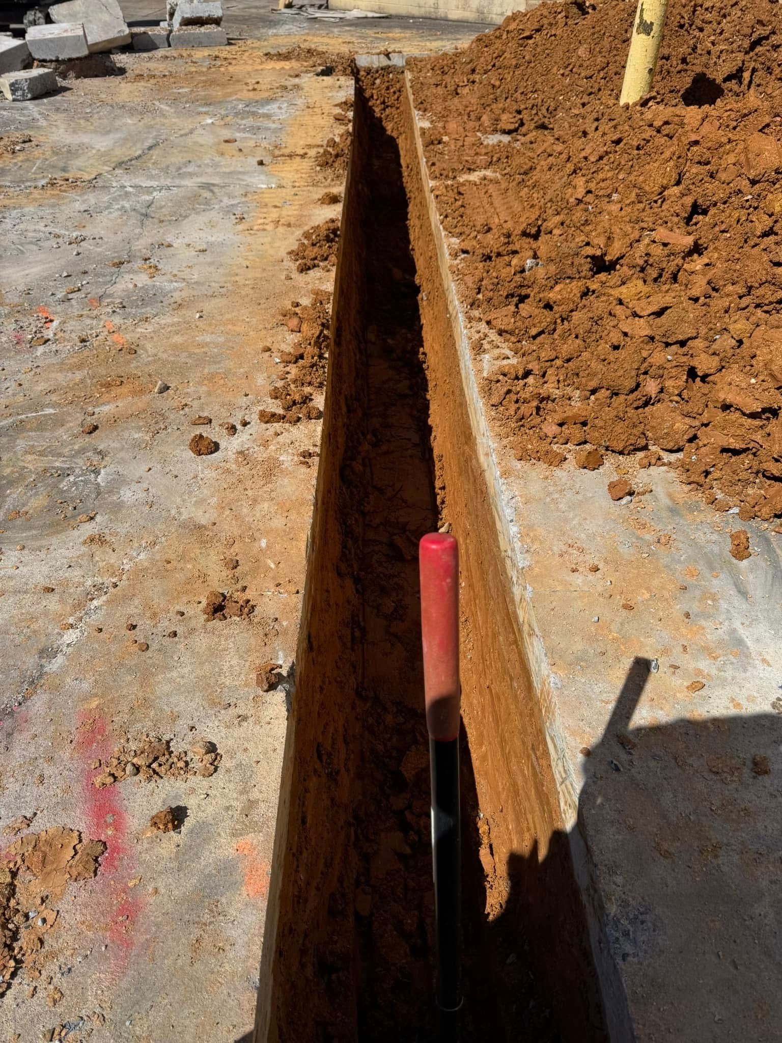 Narrow trench cut into dirt beside a concrete surface, with a red marker post at the bottom.