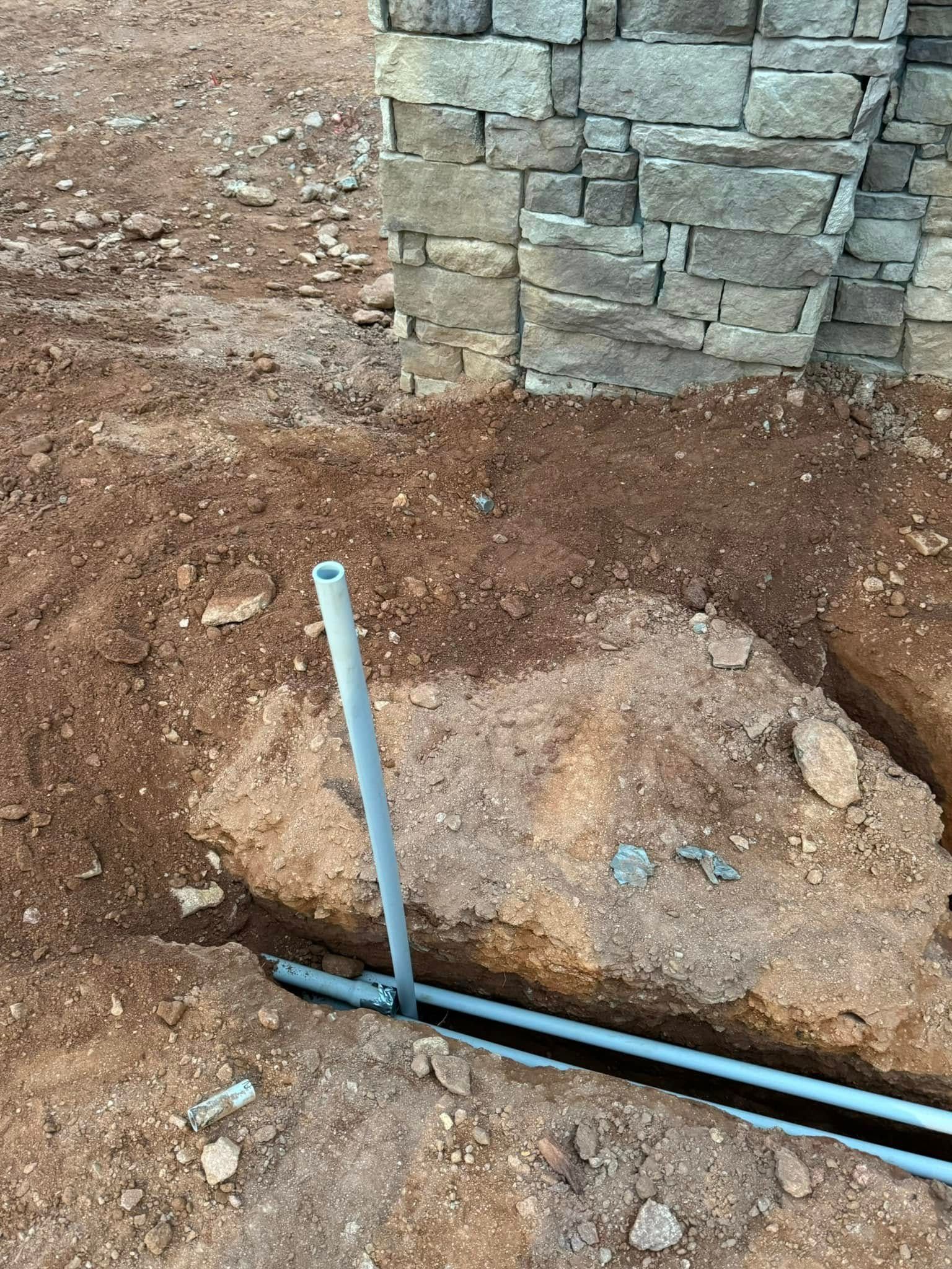 Construction site trench beside stone wall with blue pipe and white marker stake