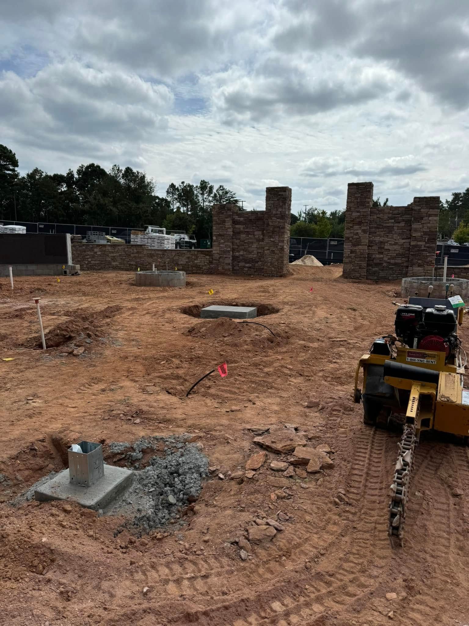 Construction site with brick columns, dirt ground, cloudy sky, and a yellow machine on the right