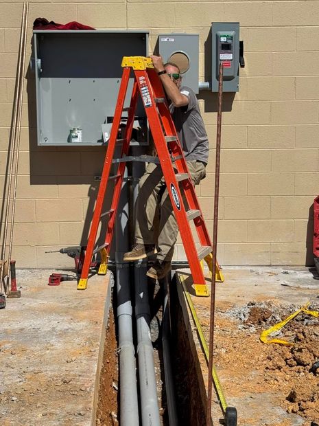 Worker on orange ladder servicing electrical panels beside an open trench outdoors.