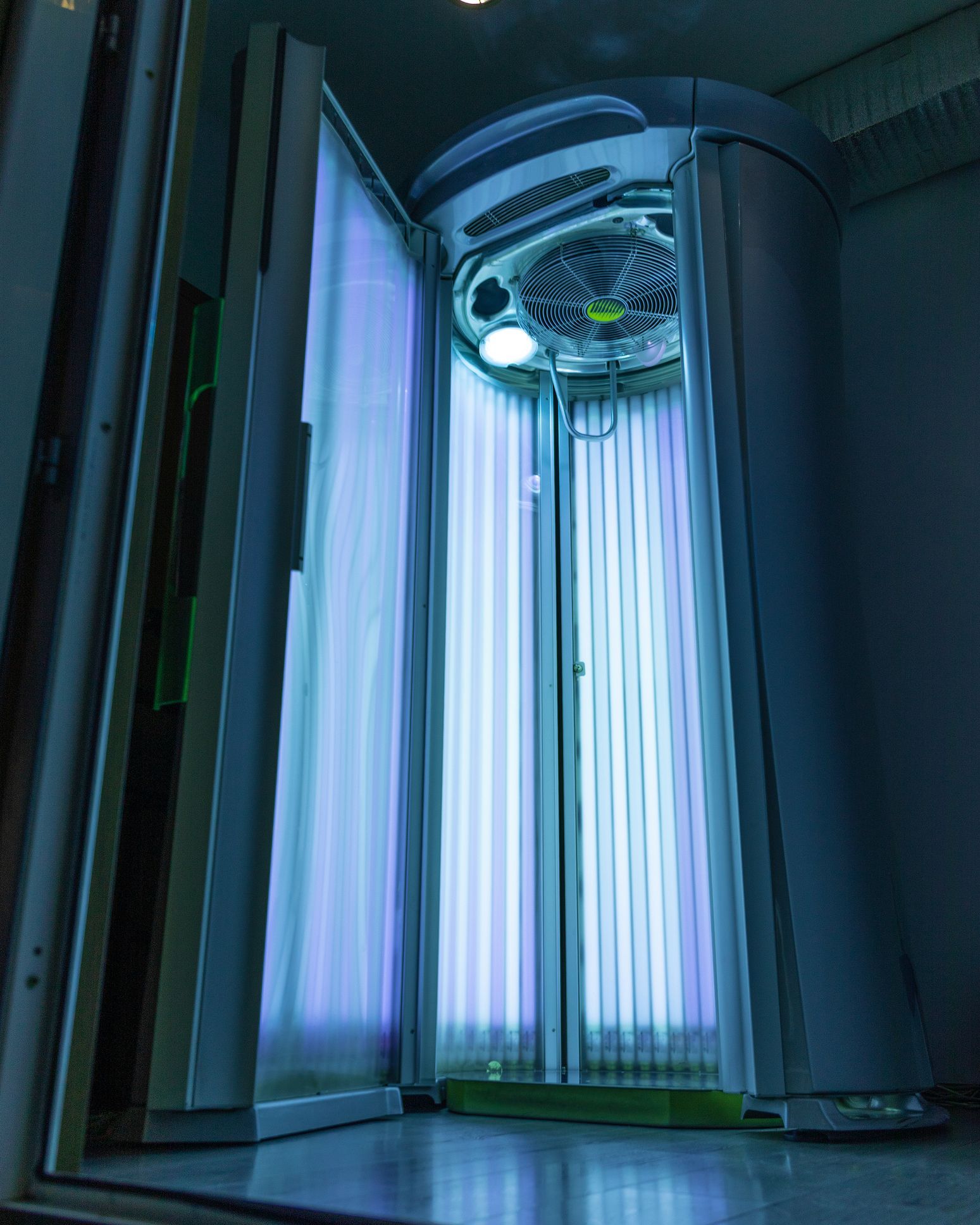 A tanning bed with open door, emitting bright blue light. The interior is dark, and the machine is silver.