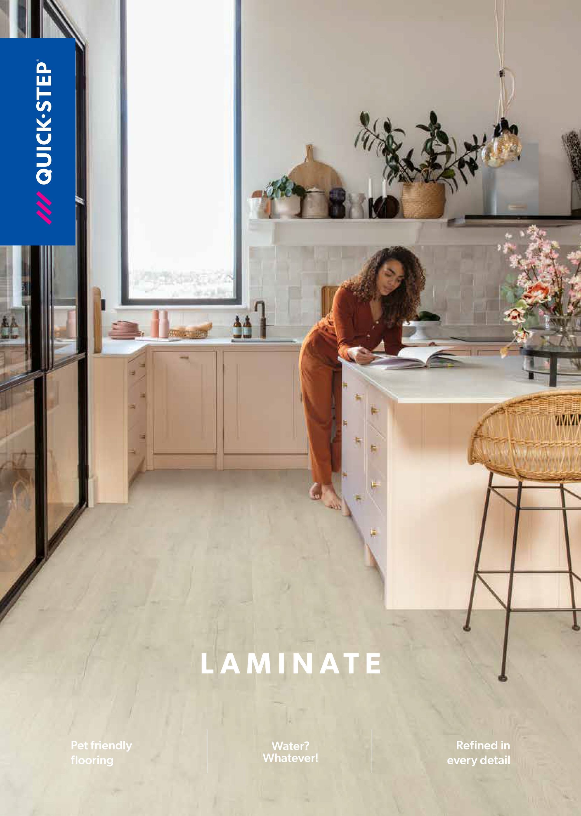 A kitchen with laminate flooring. Woman reading at a counter, surrounded by light pink cabinets and a large window.