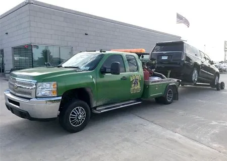 Green tow truck hauling a black SUV outside a building with an American flag