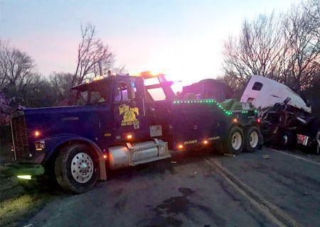 Tow truck at accident scene, night. Truck pulling damaged semi-truck, flashing lights on. Roadside setting, trees.