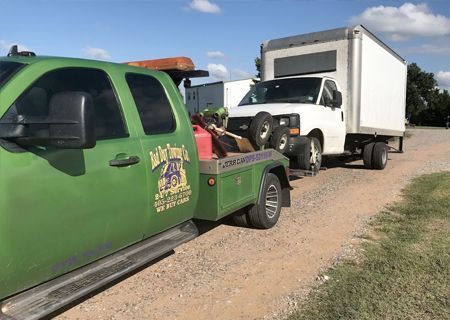 Green tow truck towing a white box truck on a dirt road.