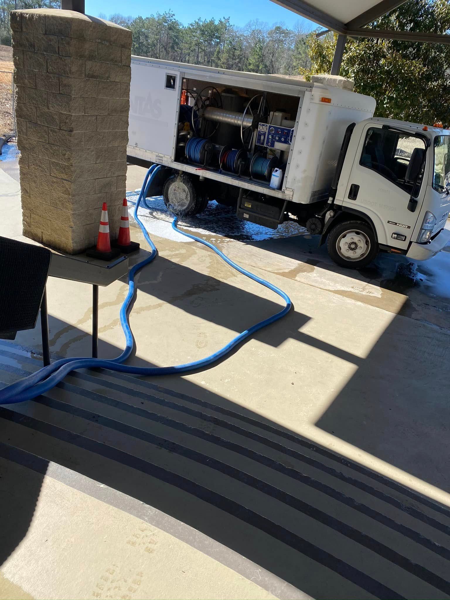 White utility truck with open side compartment parked by a building, blue hose on the ground.