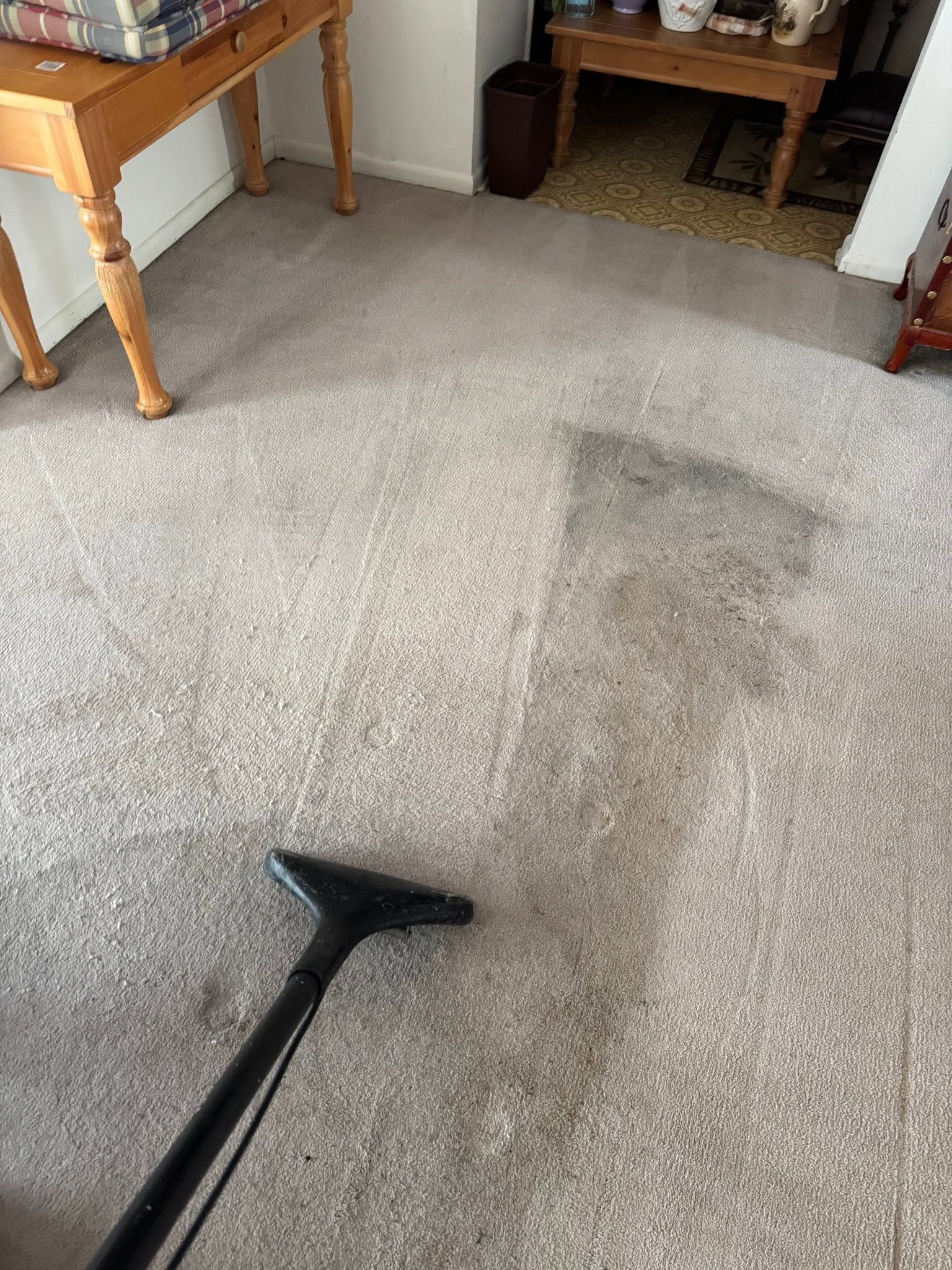 Vacuuming a beige carpet in a room with wooden tables and a chair.