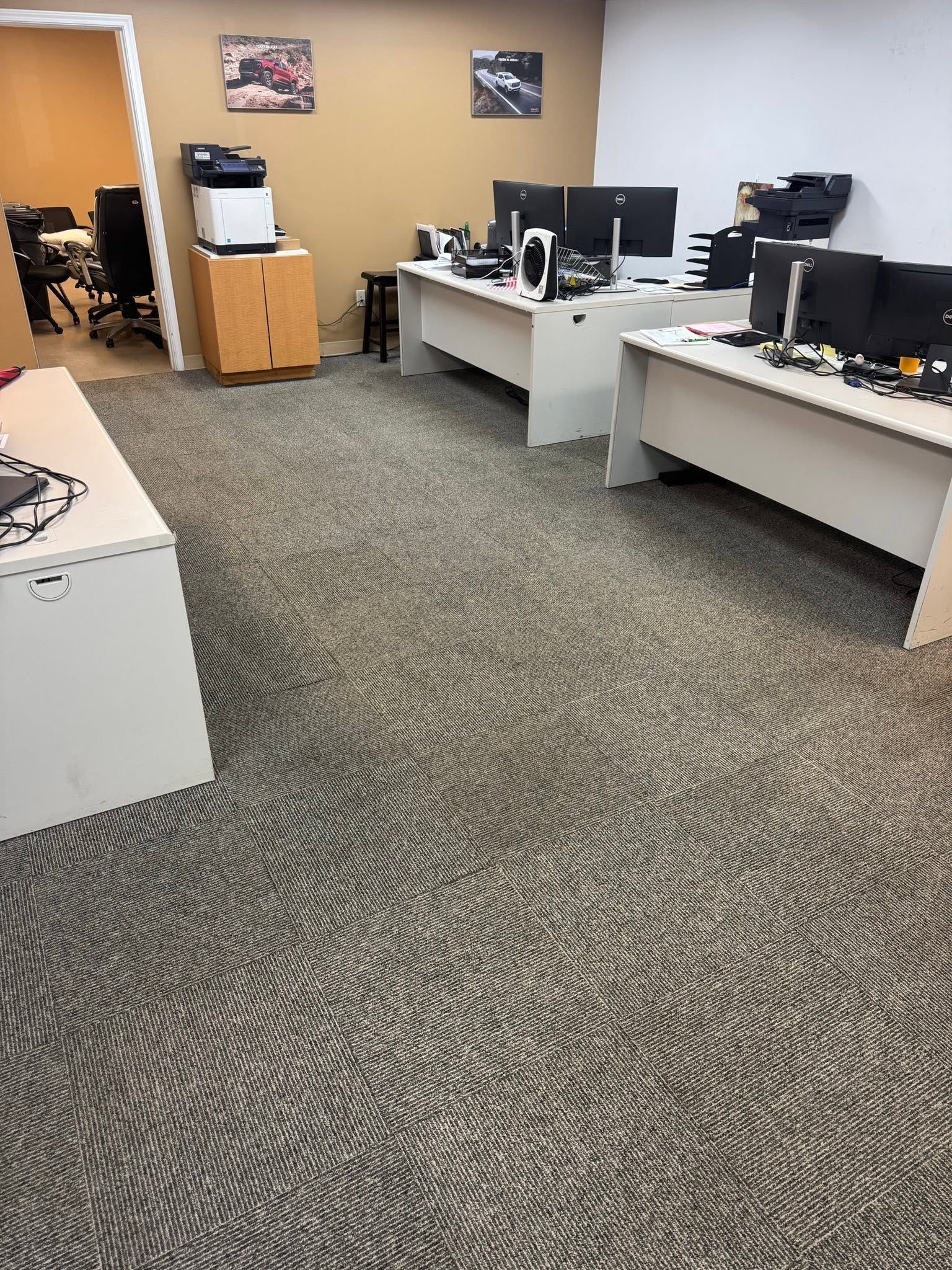 Office with rows of desks, computers, and beige walls, viewed from a carpeted aisle.