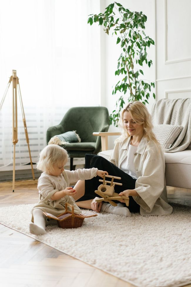 Woman and child playing on a rug in a bright living room with a toy airplane and plants.